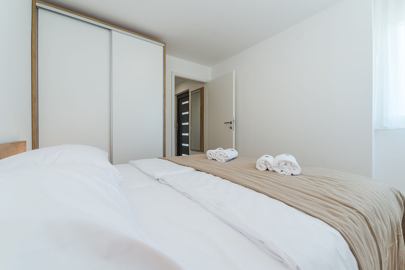 Svijetla i moderna spavaća soba, karakteristična za apartmane Vir, s uredno namještenim krevetom, svježom bijelom posteljinom, mekanim bež prekrivačem i smotanim ručnicima, pored prostranog bijelog kliznog ormara