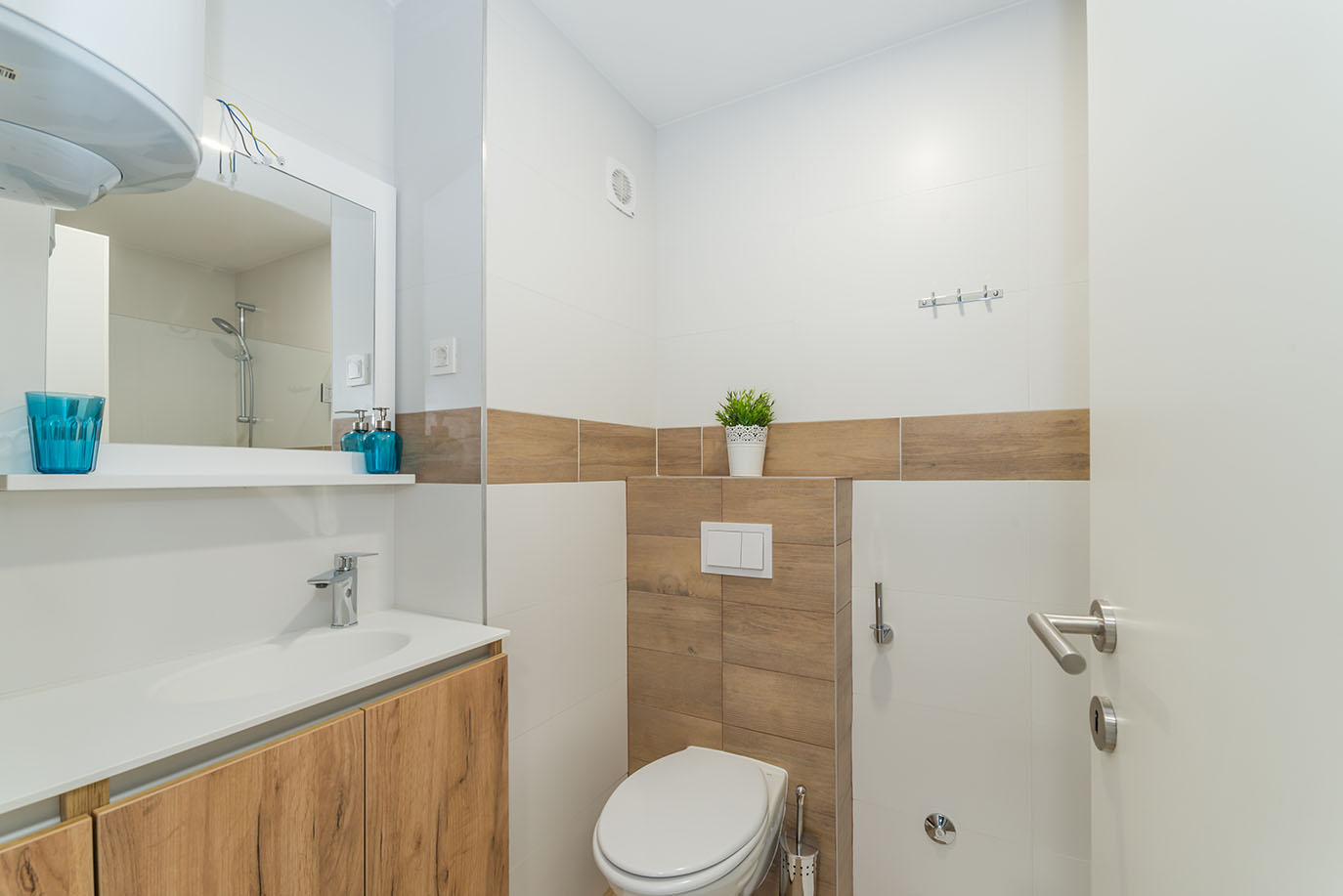 Interijer moderne kupaonice s bijelim ormarićem za umivaonik, drvenim elementima, WC školjkom i pločicama u bijeloj i drvenoj boji, tipično za apartmane Vir