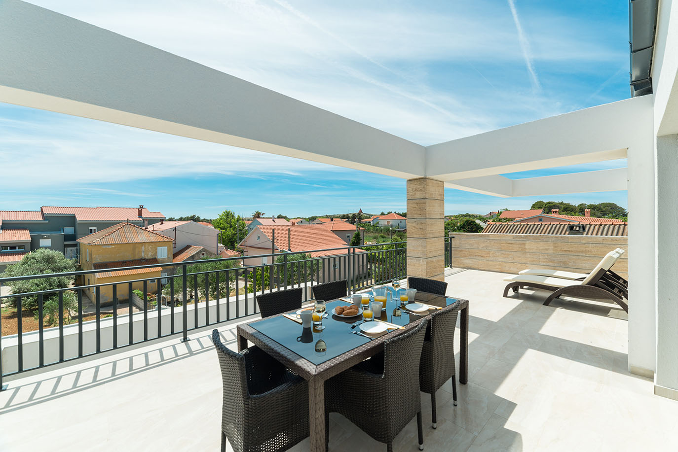 Prostrana krovna terasa s postavljenim stolom za doručak, udobnim ležaljkama i panoramskim pogledom na mjesto s crvenim krovovima, idealna za goste apartmana Vir