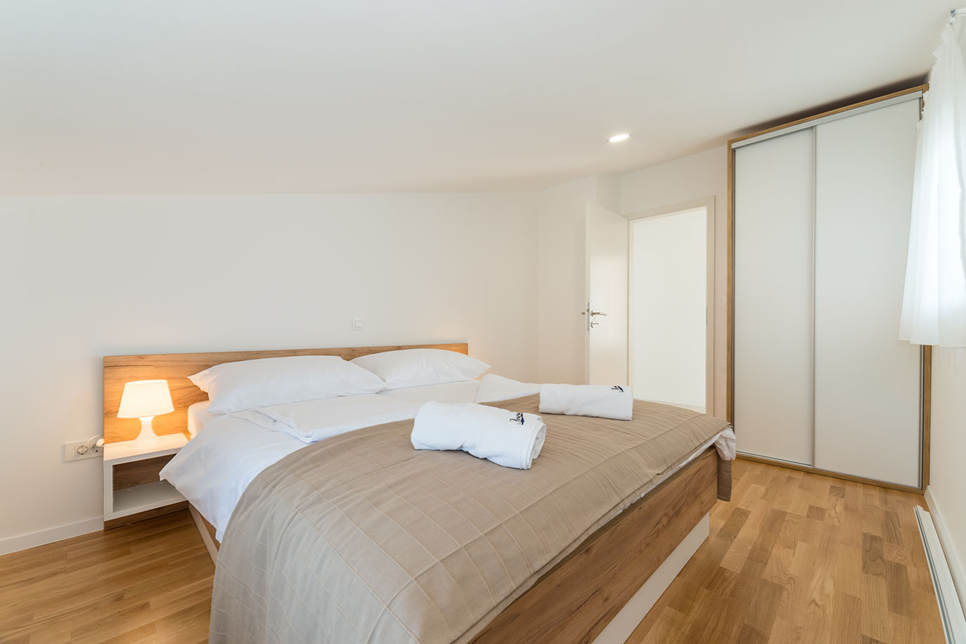 Svijetla i moderna spavaća soba s velikim krevetom s drvenim uzglavljem, bijelom posteljinom, dvama smotanim bijelim ručnicima, noćnom lampom i bijelim kliznim ormarom, karakteristična za kvalitetne apartmane Vir