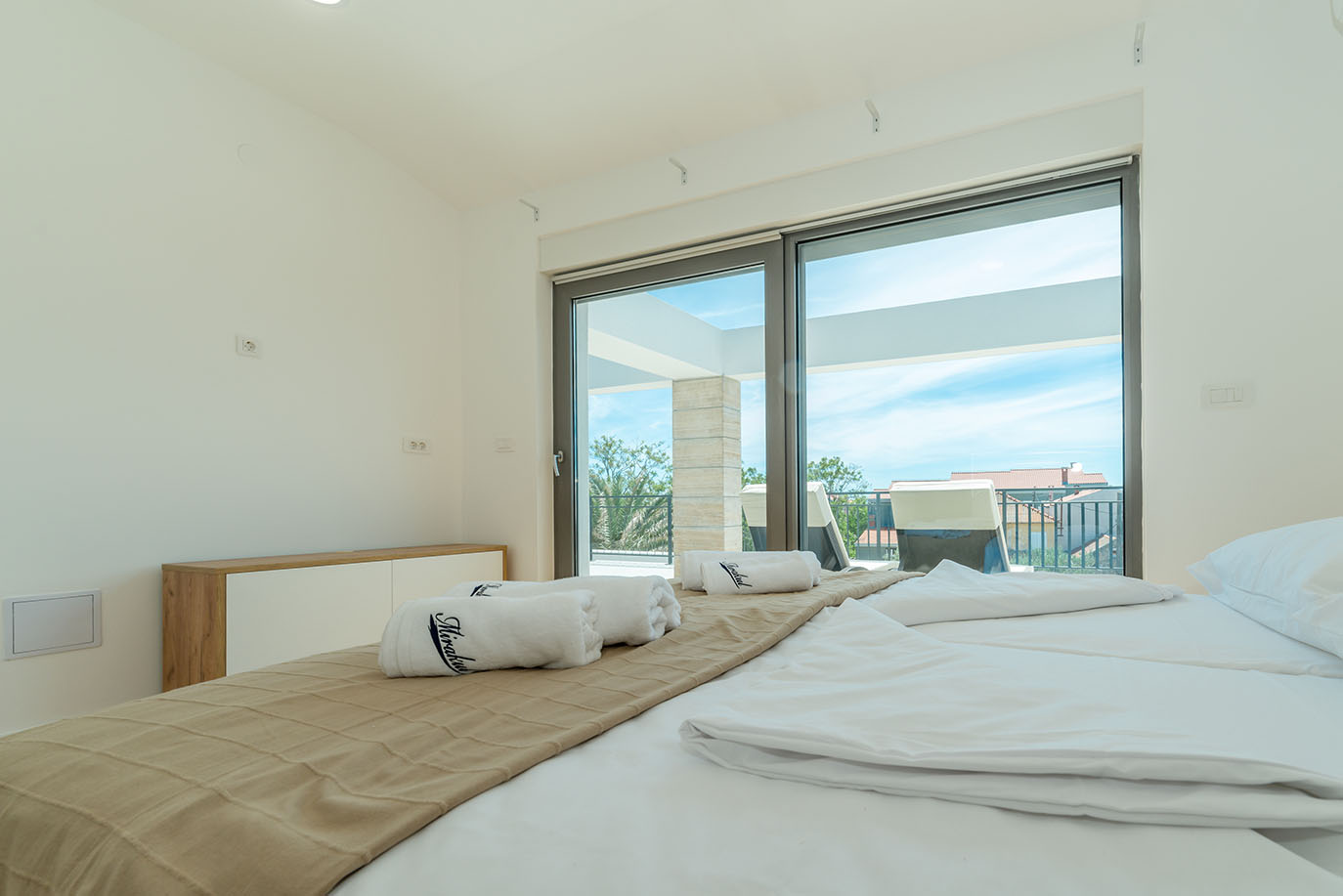 Svijetla i moderna spavaća soba u Villi Mirakul s urednim krevetom, bijelom posteljinom, brendiranim ručnicima i velikim staklenim vratima koja vode na sunčani balkon