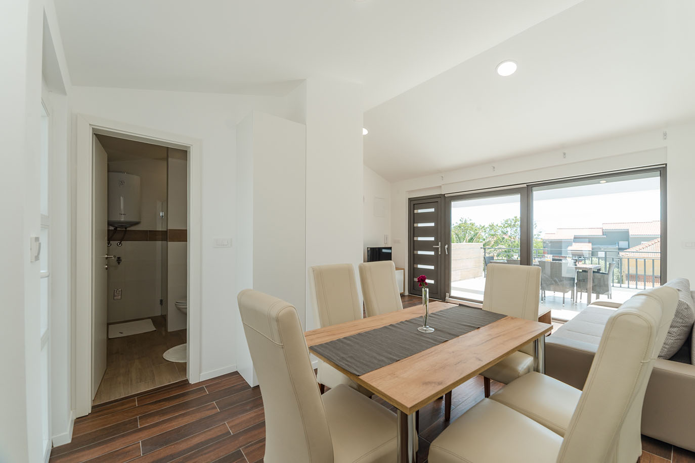 Svijetli i ugodni otvoreni dnevni boravak i blagovaonica u apartmanima Vir, namješteni modernim drvenim stolom, krem stolicama, udobnom sofom i velikim kliznim vratima koja vode na sunčanu vanjsku terasu